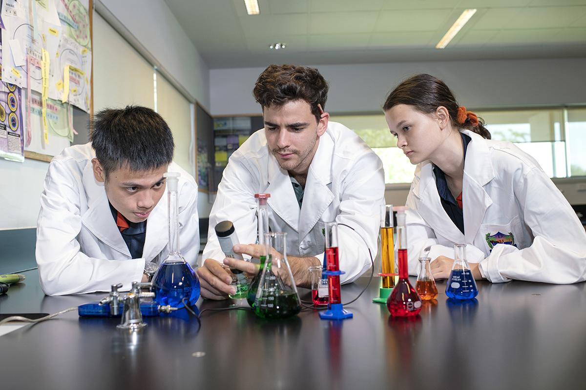 Three students in white lab coats looking beakers and equipment with coloured fluid in them.